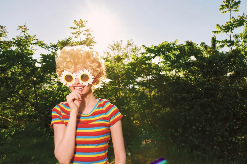 Cheerful woman outdoors, striped shirt and sunflower sunglasses, enjoying a sunny breeze among lush trees, bright colors, relaxed pose, candid lifestyle moment in nature.