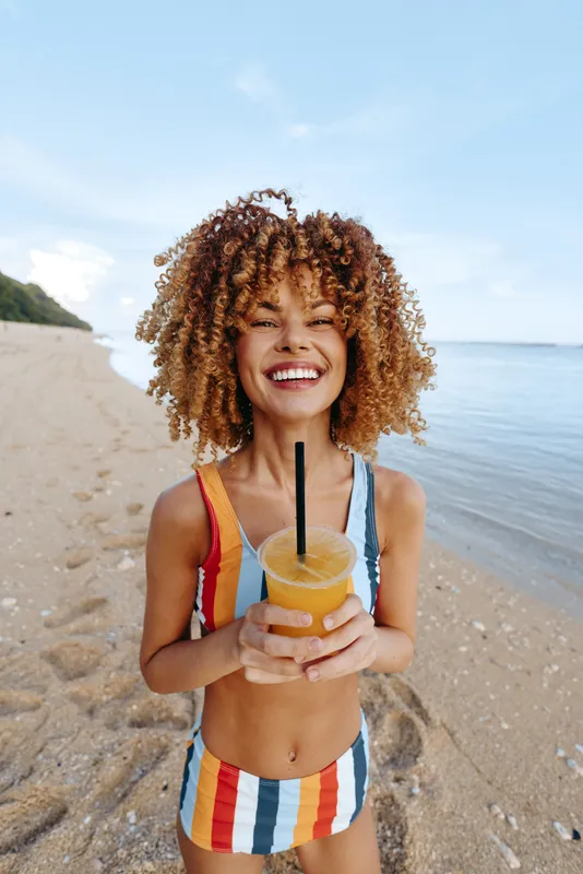 Smiling young woman with curly hair on a bright sunny beach close up portrait holds a cold juice drink with a straw, wearing a striped swimsuit, summer vacation leisure mood, calm ocean background