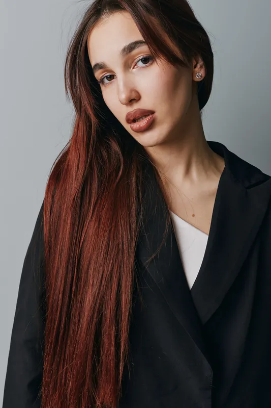 Woman with long straight brown hair wearing black blazer and white top posing against gray background. Portrait of young female model with natural makeup and serious expression.