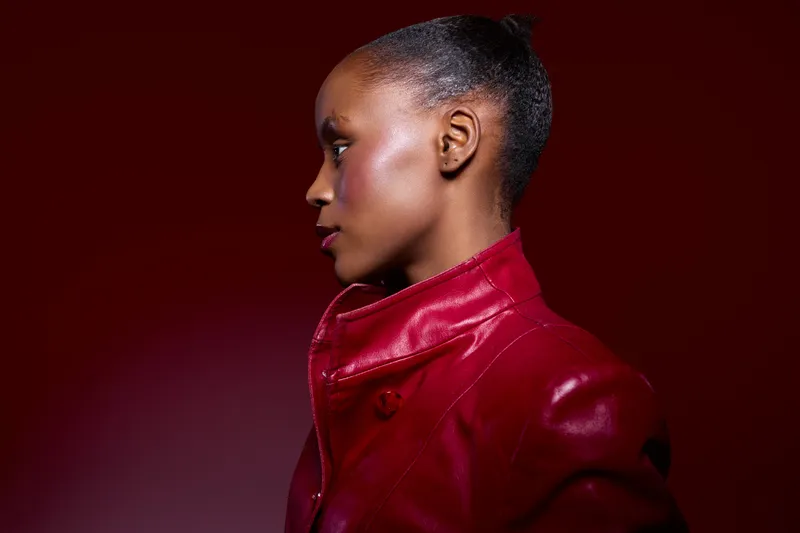 Profile of a young black woman with dark skin wearing a red leather jacket against a red background. Studio portrait with dramatic lighting highlights her smooth skin and natural hairstyle.