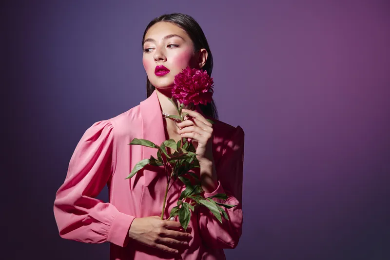 Elegant woman wearing pink with bold red lipstick poses with a deep red flower in front of purple background. Concept for beauty fashion creative photo session and makeup art.
