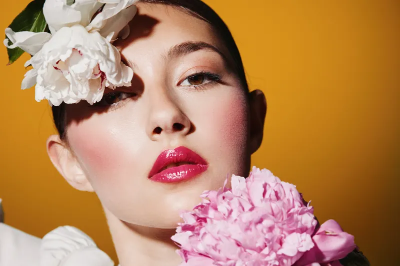 Close-up portrait of a young woman with glossy pink lips and floral accessories on her face. Bright artistic makeup on warm orange background for beauty creative fashion.