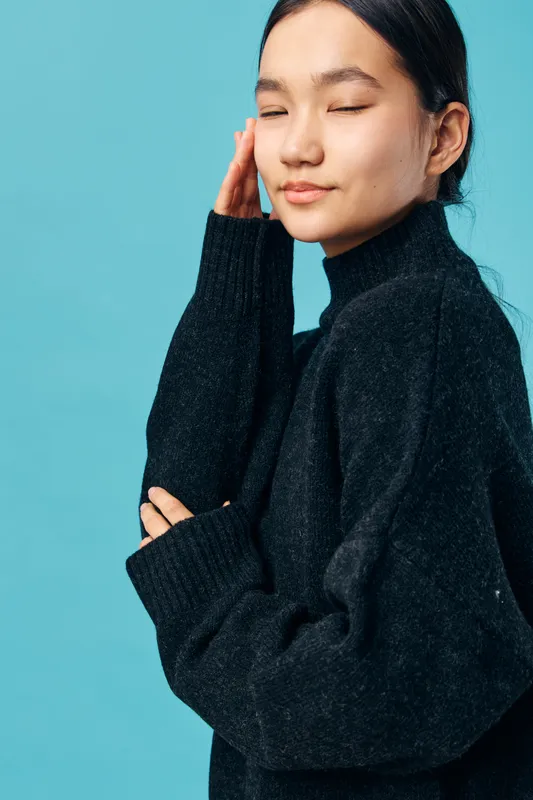 Young woman in black sweater with eyes closed and hand touching face, posing peacefully against blue background in studio portrait for fashion and beauty concepts.