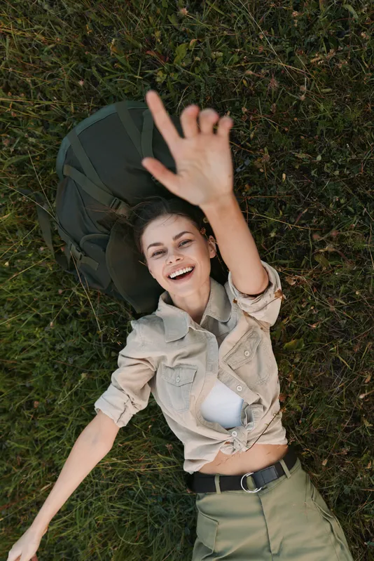 Young woman lies on green grass outdoors, smiling and reaching up beside a backpack, capturing a carefree lifestyle moment.