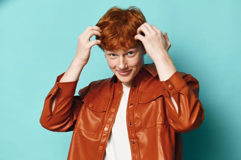 Young man with red hair wearing brown leather jacket and white shirt smiling while touching hair against blue background in studio portrait.