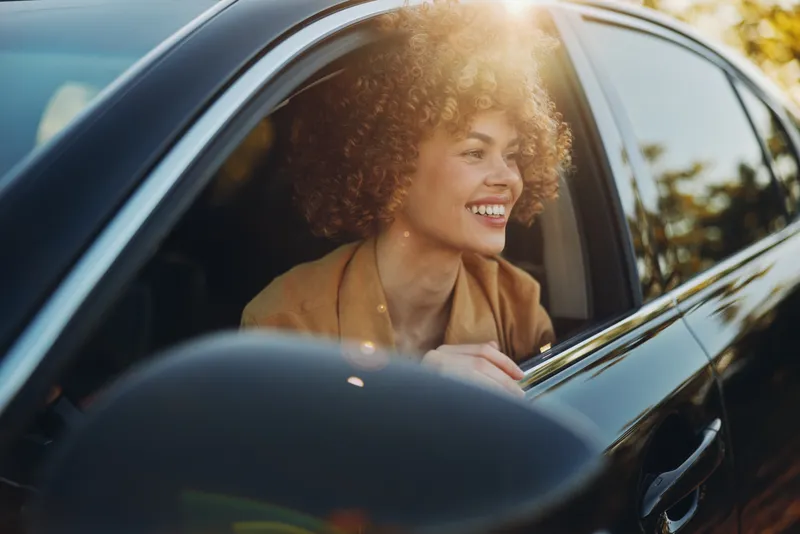 Woman smiles from car window, enjoying a carefree lifestyle, warm retro film tones, candid moment inside a stylish vehicle, natural light enhances joyful mood evokes freedom, travel, sunset glow and