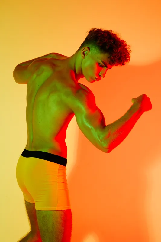 Muscular man posing shirtless in colorful lighting, showing strong back and arm muscles with yellow shorts in studio for fitness and bodybuilding concept.