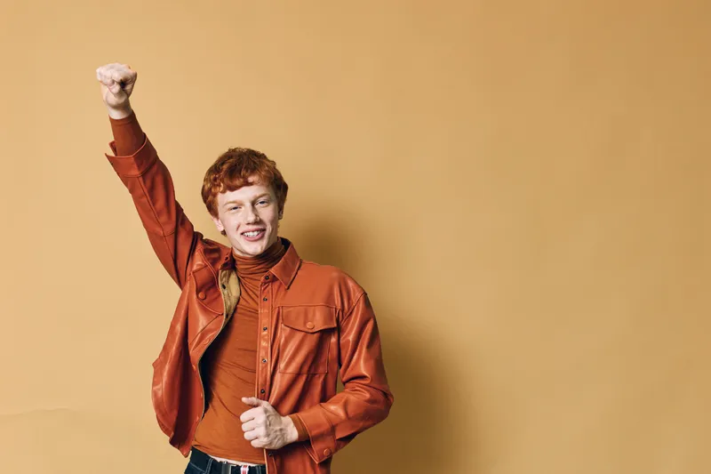 Young man with red hair wearing orange jacket and turtleneck stands with raised fist against beige background smiling confidently with positive gesture.