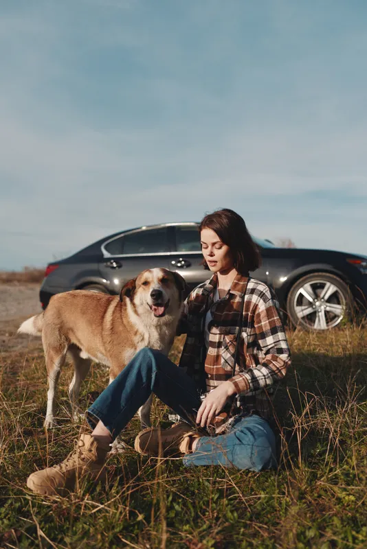 Woman wearing a plaid shirt and jeans sits on tall grass with a dog beside her, car in the background, enjoying an outdoor casual moment in a sunny field.