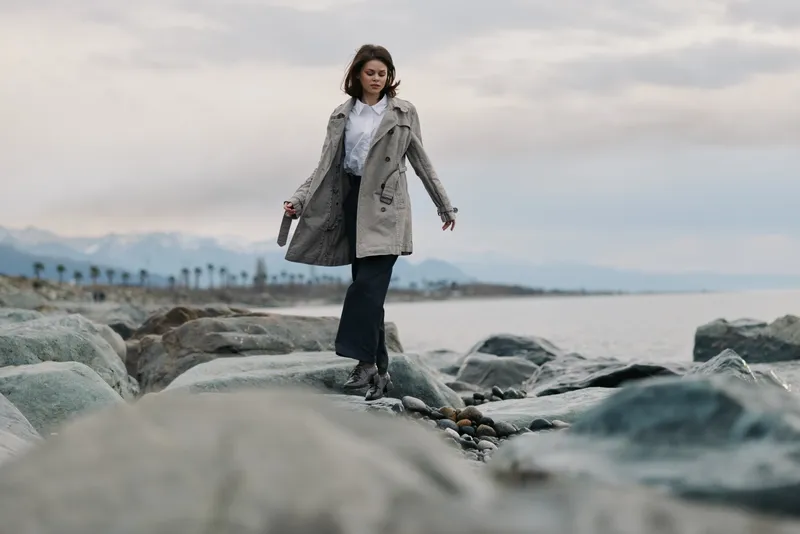 Young woman in coat walks carefully on rocky beach near sea with cloudy sky. Concept of nature, travel, exploration, and outdoor adventure in cool weather.