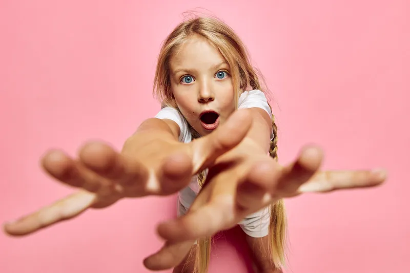 Surprised girl with long blonde hair reaches toward the camera with open mouth on a pink background. Studio portrait with enthusiastic expression and playful pose. Concept of childhood.