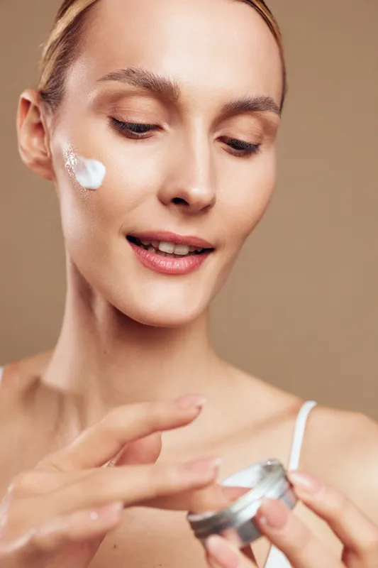 Young woman applying face cream with gentle smile soft natural skin closeup beige background beauty care concept moisturizing treatment daily routine