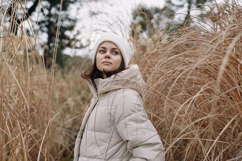 Woman in a hooded winter coat stands in tall dried grass, outdoors among nature, a calm portrait capturing chilly atmosphere and soft natural light