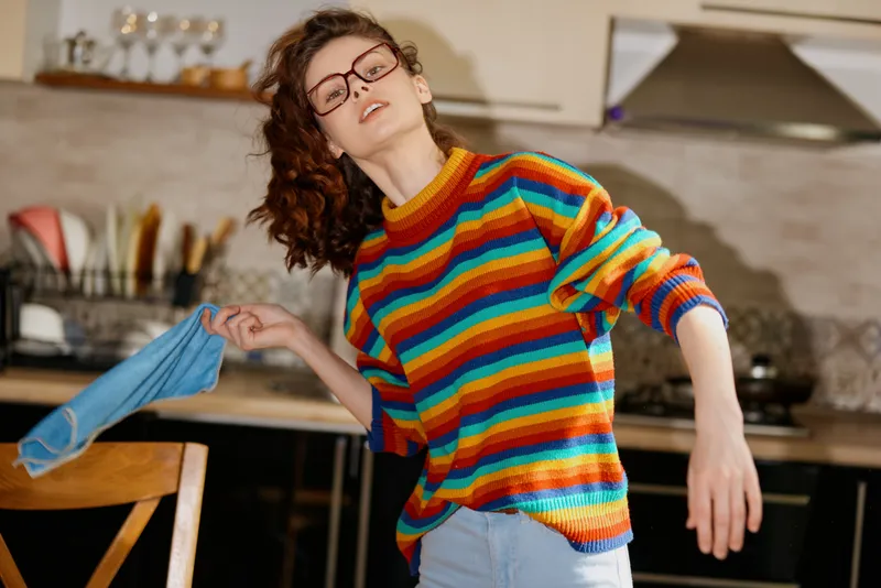 Candid woman with colorful striped sweater in a bright kitchen, leaning and reaching with a cloth. Lifestyle scene capturing home, daily activity, and cheerful energy.
