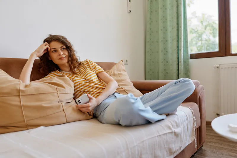 A relaxed woman lounges on a sofa at home, wearing a striped shirt and jeans, soft natural light highlights comfortable living. Concept: home comfort and casual relaxation.