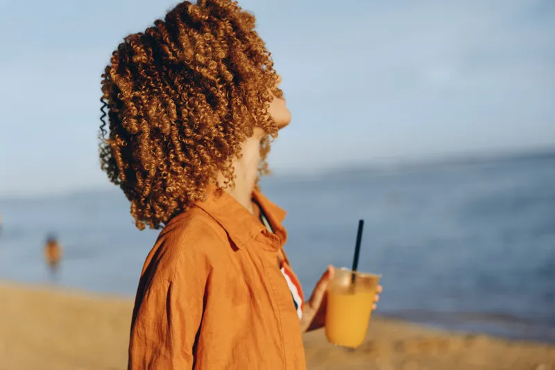 A young woman with curly hair wears an orange rust shirt and holds a drink with a straw as she stands on a sunny beach, gazing toward the calm sea in the background.