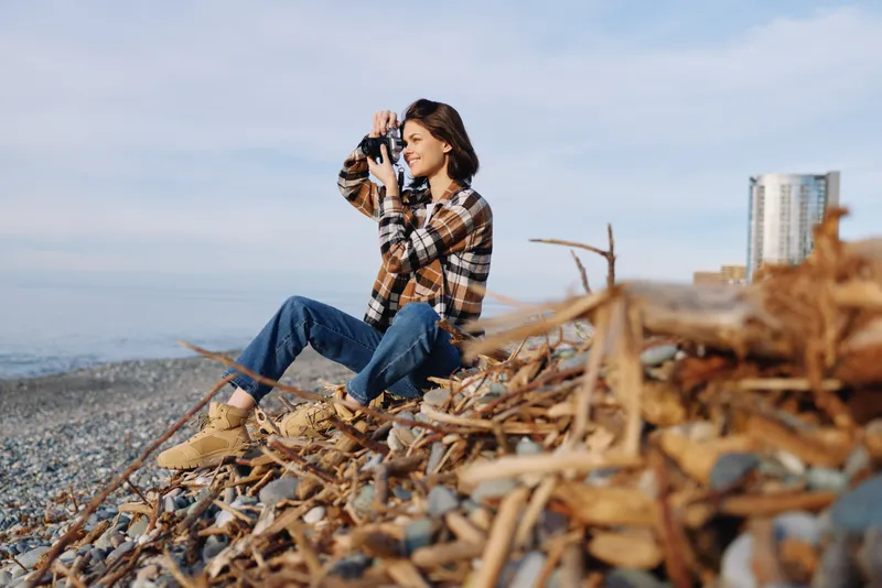 Woman photographer on a rocky beach wearing a plaid shirt and jeans, capturing the horizon with a handheld camera among driftwood and coastal breeze