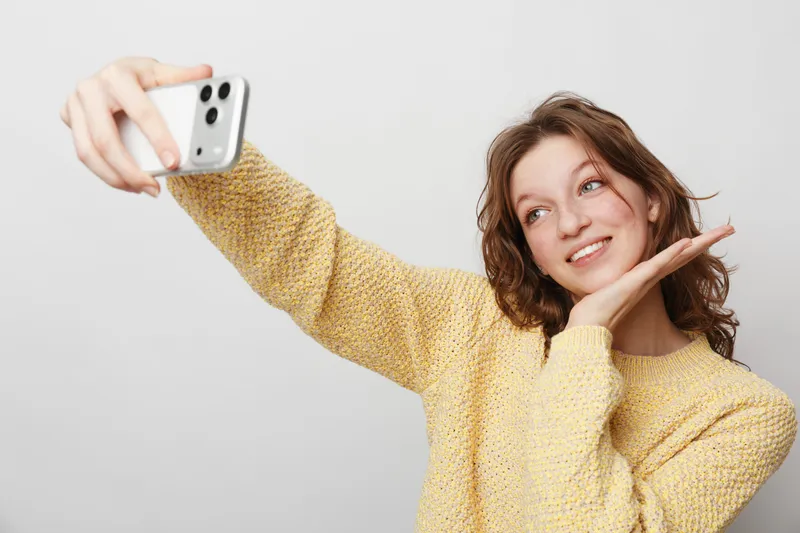 Young woman in yellow sweater takes selfie with smartphone, smiling and posing with hand near face. Bright studio background, casual style and positive mood concept.