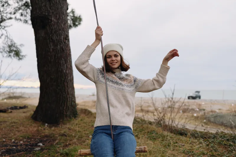 Girl on a wooden swing among trees in a chilly outdoors setting, wearing a cozy sweater and beanie, enjoying a playful moment under gray skies.