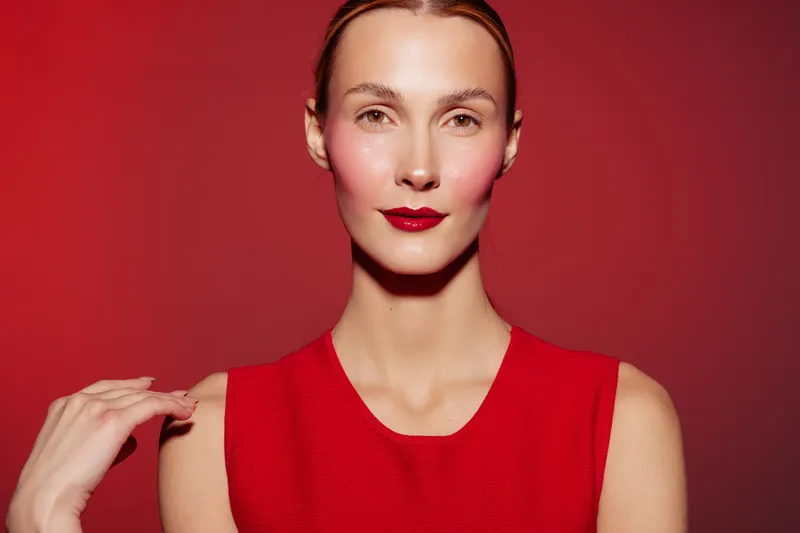Portrait of confident woman with red lipstick and sleeveless red dress on red background. Studio lighting highlights natural skin texture and elegant facial features. Fashion and beauty concept.