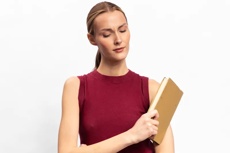 Young woman in red sleeveless top holds a closed book, looking down with a thoughtful expression, isolated on white background. Concept of reading, education, and contemplation.