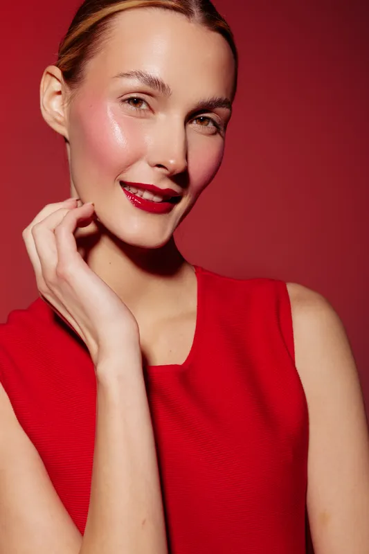Smiling woman with shiny skin and red lipstick wears red sleeveless top against red background. Concept of beauty, makeup, fashion, and confident female portrait.