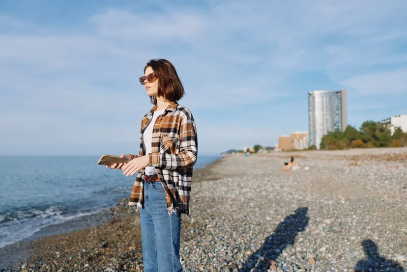 Casual woman by the seaside holding a notebook, wearing sunglasses and a plaid shirt, jeans and a calm pose as waves roll on a pebbled beach under a blue sky.