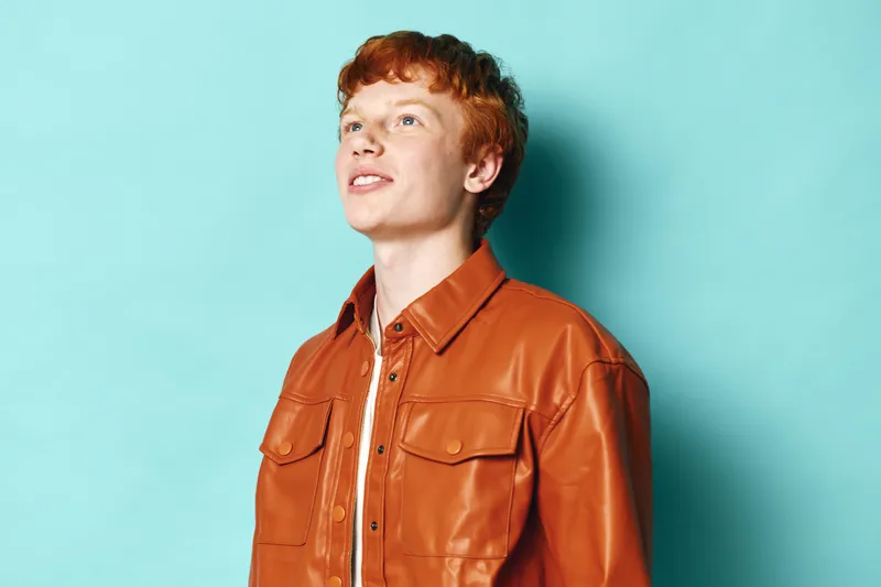 Young man with red hair wearing brown leather jacket looks upward with confident expression against blue background. Portrait of male model in casual stylish outerwear indoors.