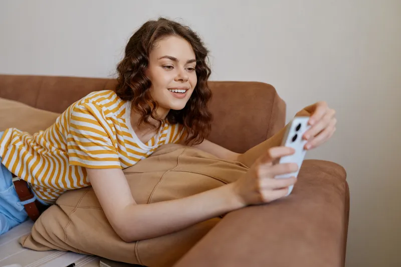 A young woman relaxes on a cozy sofa at home, smiling softly as she scrolls through her smartphone, wrapped in a striped t shirt, with warm natural light filling the room.