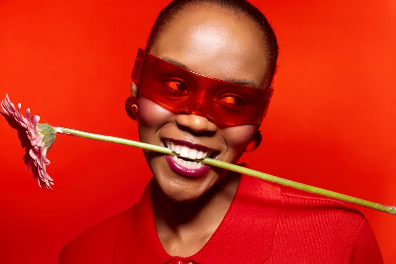 Smiling dark skinned African woman wearing red sunglasses and red outfit holds pink flower in mouth. Bright red background creates bold fashion concept with joyful and confident mood.