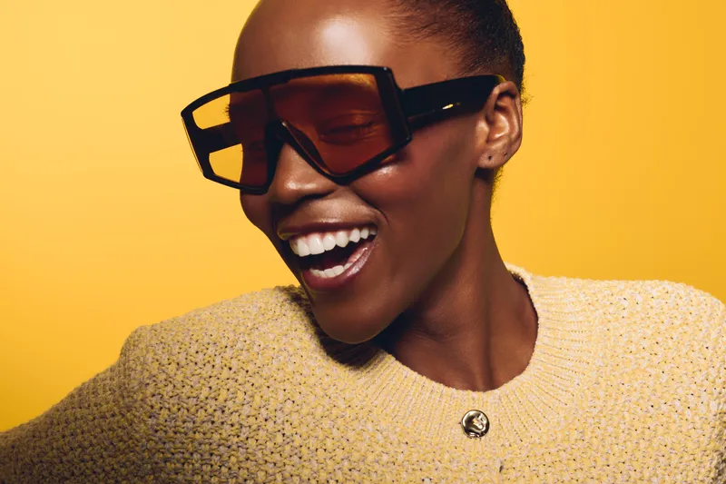 Smiling African American woman with dark skin wearing large sunglasses and yellow sweater poses against yellow background. Concept of fashion, style, joy, and modern beauty.