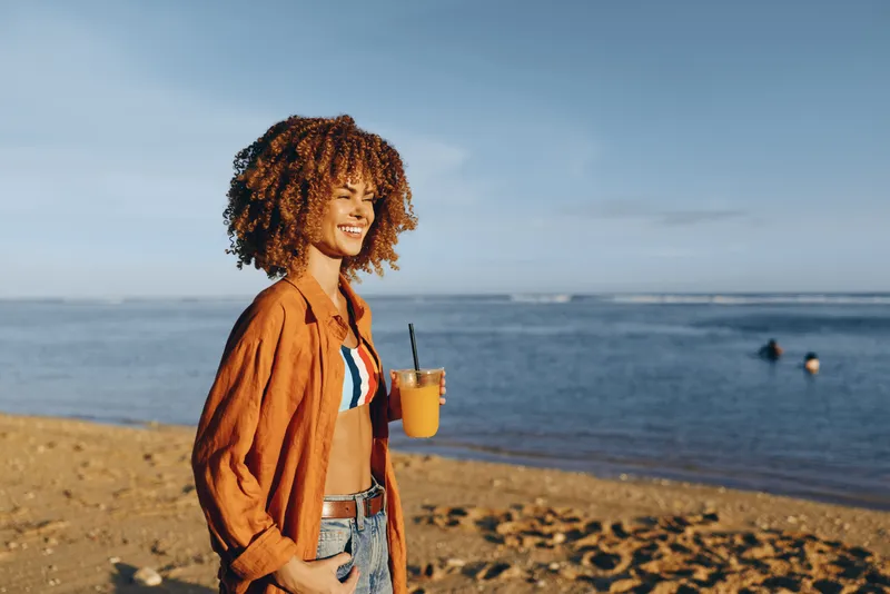 Young woman standing on beach by ocean, curly hair in wind, casual jacket and shorts, holding a refreshing drink, smiling in warm sunlight, summer vacation outdoors, calm seaside waves.