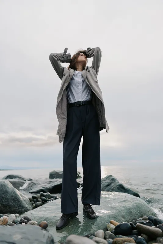 A young woman stands on rocky shore wearing a long coat and wide pants. She poses with hands on her head against a cloudy sky and calm sea background. Fashion and nature concept.