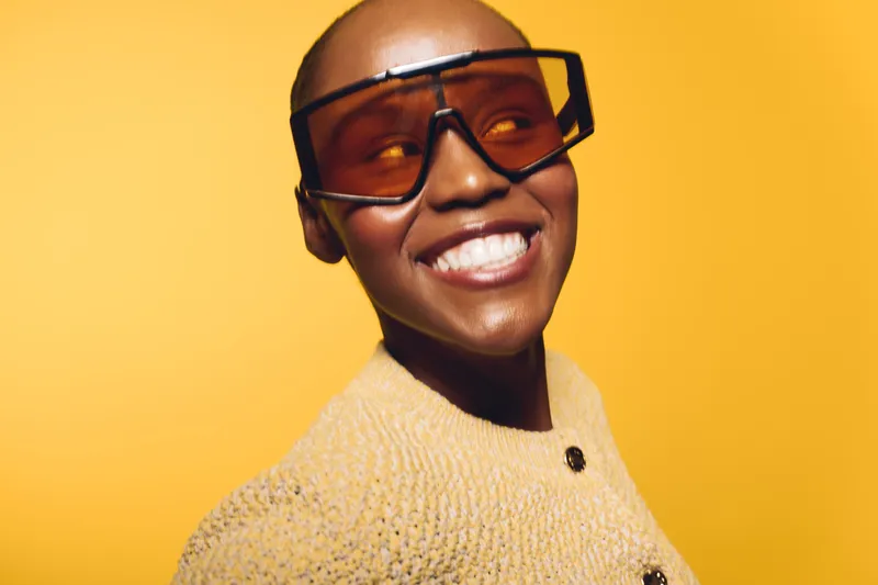 Smiling black woman with dark skin wearing oversized sunglasses and a textured yellow sweater stands against a bright yellow background. Concept of fashion, confidence, and joy.