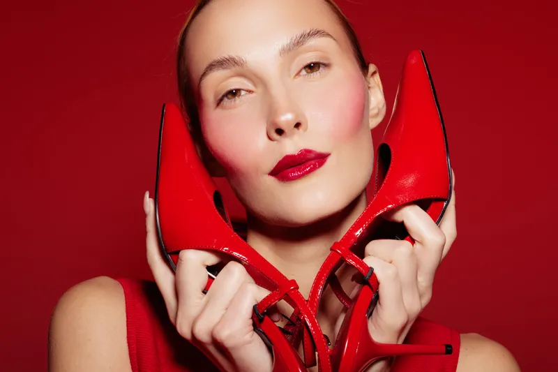 Close-up of a stylish woman holding red high heels near her face against a red background. Concept of fashion, beauty, glamour, and bold color contrast in studio lighting.