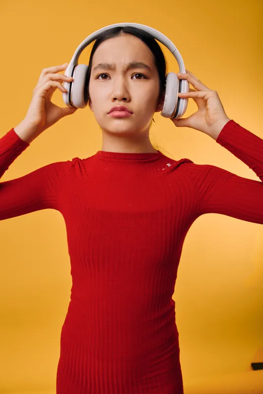 Young woman in red sweater wearing wireless headphones adjusting them with both hands while standing against yellow background. Concentrated expression and casual style with audio technology.