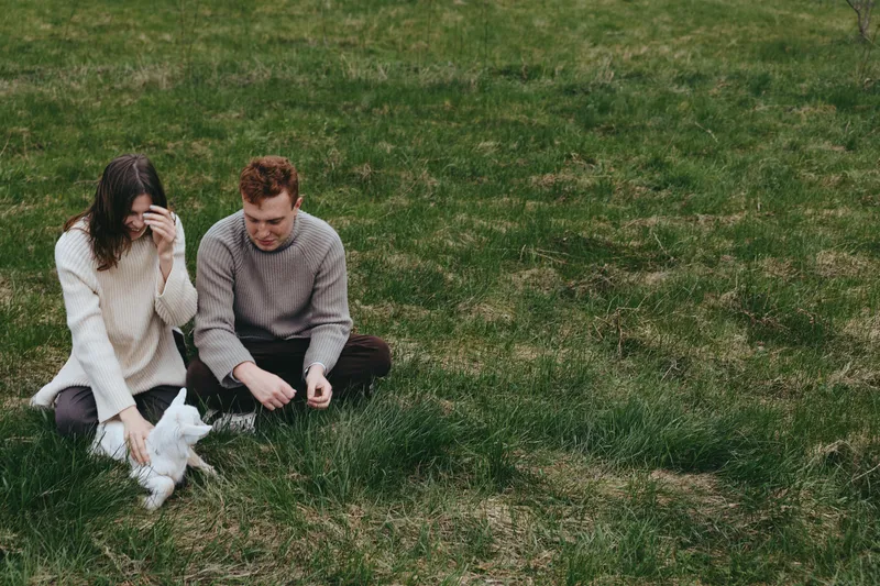 Couple sits on grass outdoors sharing a playful moment with a goat kid nearby, creating a warm lifestyle scene focused on companionship and gentle animal interaction in soft light. Cozy.