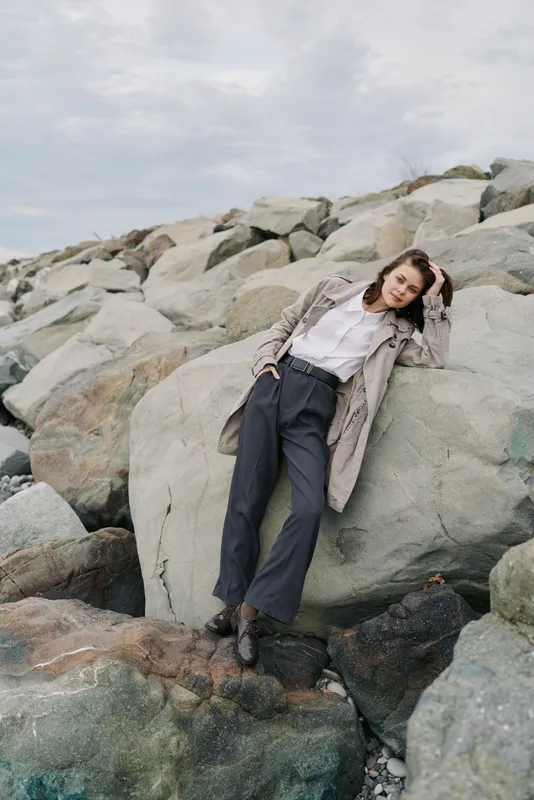 Young woman in stylish outfit leans on large rocks near the coast. Overcast sky and natural stone background create a calm and contemplative mood for fashion and lifestyle concept.