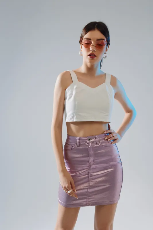 Young woman in a white crop top and shiny purple skirt poses confidently wearing pink sunglasses. Studio shot with soft lighting. Concept of beauty, fashion, and style.