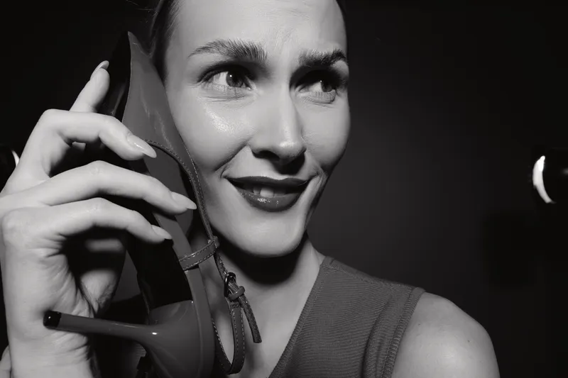 Close-up of a smiling Caucasian woman with light skin holding a high heel shoe to her ear as if it were a phone, black and white photo with dramatic lighting and dark background.