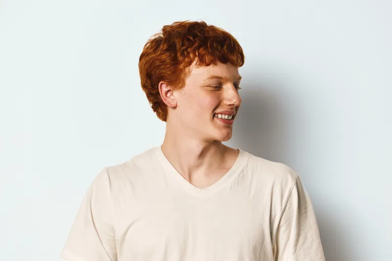 Young man with red hair and freckles smiling and looking to side wearing light beige t shirt on white background studio portrait.