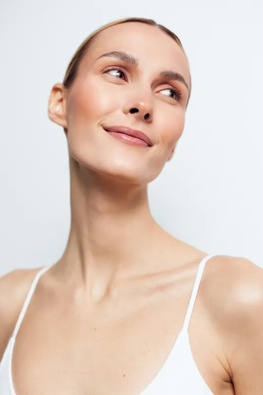 Close up portrait of a smiling woman in a white top, with soft lighting and glowing skin, captured in a minimalist studio setting to emphasize calm confidence, natural beauty, and relaxed mood.