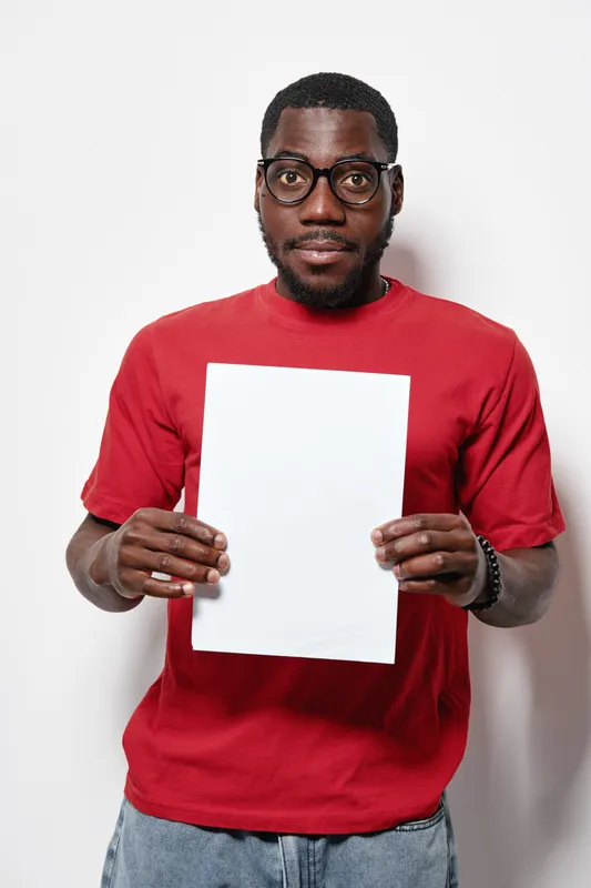 African American man with dark skin wearing glasses and red shirt holding blank white paper in front of white background for message or advertisement concept.