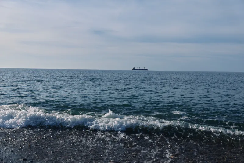 Calm sea scene with gentle waves on a pebbled shore, distant ship on the horizon and clear blue water under a light sky, capturing a tranquil coastal moment.