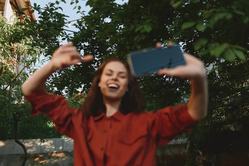 Cheerful woman outdoors taking a selfie, laughing with head tilted, sunlit moment, relaxed pose, soft breeze, natural colors, lush greenery background, candid lifestyle scene