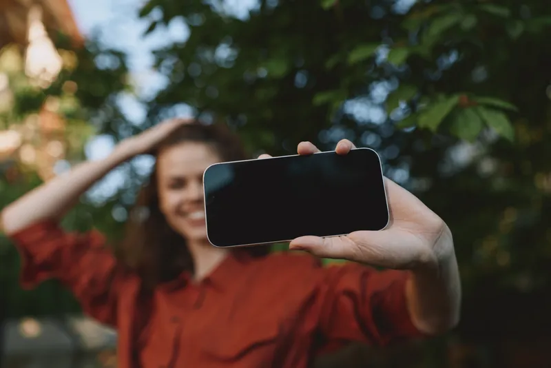 Smiling woman taking a selfie outdoors with a smartphone, bright daylight, natural greenery in the background, casual lifestyle moment, candid and lively mood