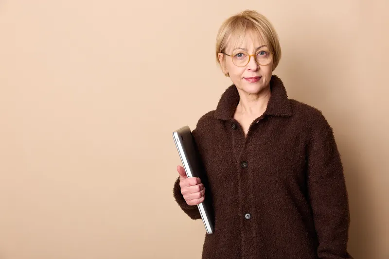 Woman elderly glasses book portrait education reading library home beige background, calm mature female in coat holding book, studio portrait with copy space, indoors learning and wisdom.