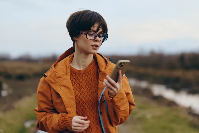 Woman outdoors in countryside, wearing orange jacket and knit sweater, looking down at smartphone, relaxed lifestyle moment, soft daylight, grassy field and blurred river background, nature