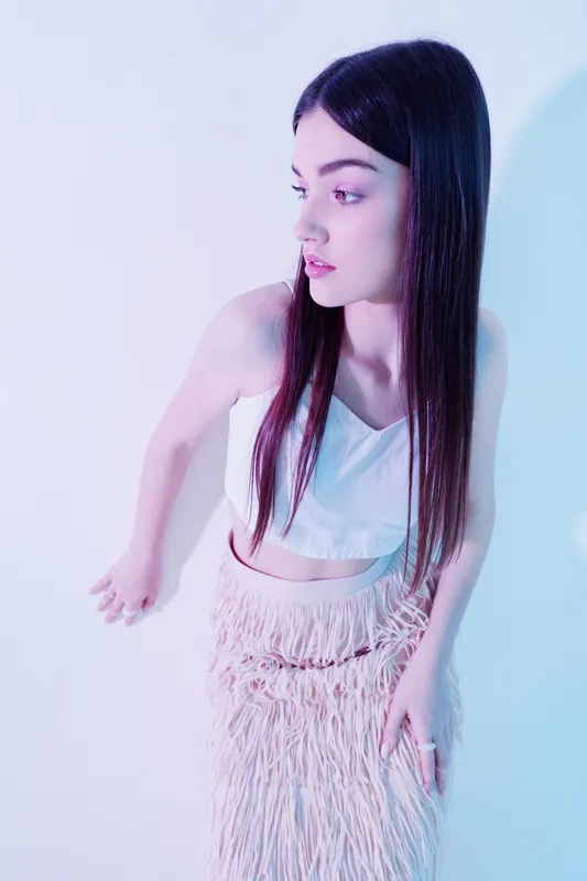 Young woman with long dark hair wearing a white crop top and textured skirt poses against a light background. Concept of woman beauty fashion and modern style.