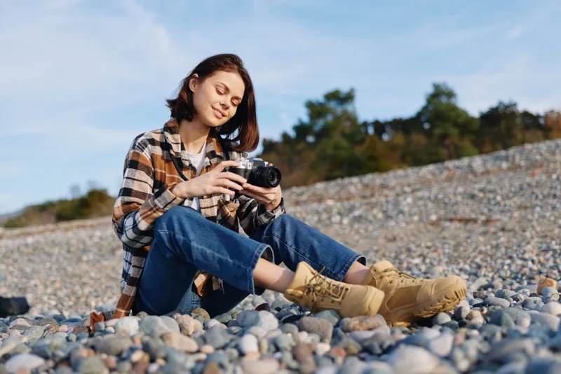 Woman photographer sitting on a pebble beach, examining a camera in daylight, wearing a plaid shirt, jeans and boots, focused on capturing natural landscape and candid outdoor moments.